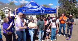 A group of people stand in front of a tent for Lutheran Family Services and smile as a ribbon has just been cut in a community garden.