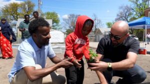 A small child wearing a red hoodie plants a vegetable with the assistance of two men on each side.