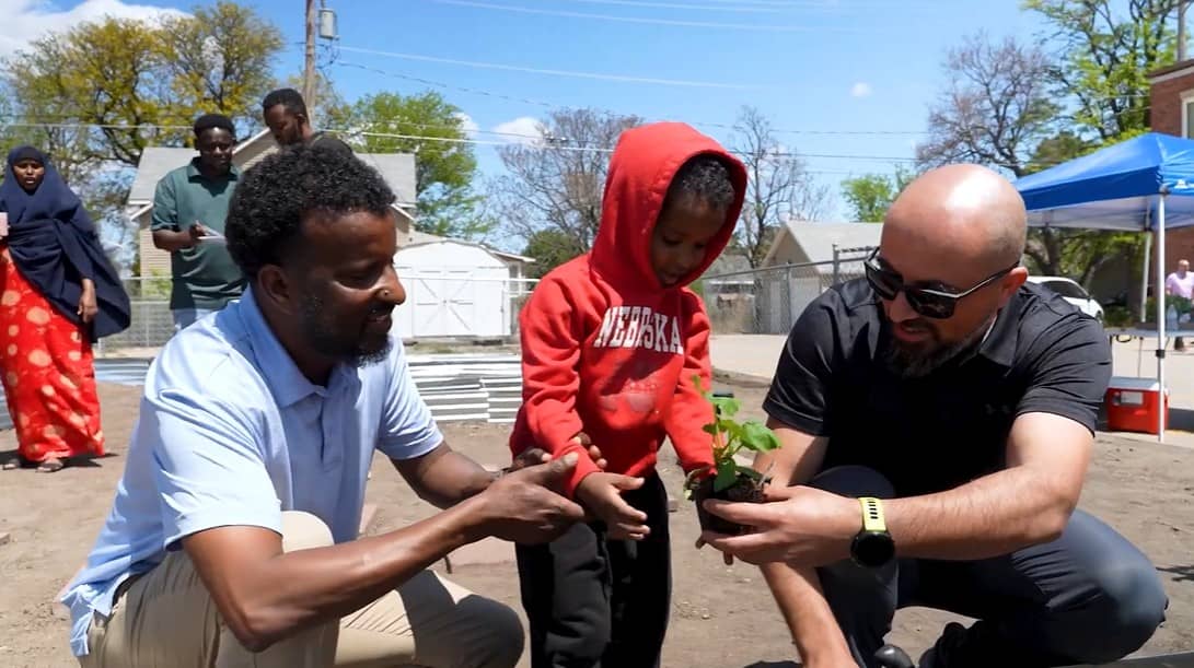 A small child wearing a red hoodie plants a vegetable with the assistance of two men on each side.