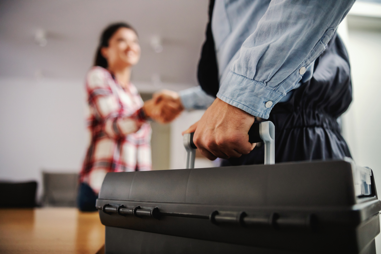 A home repair man holds a tool box and shakes hands with a female homeowner wearing a flannel shirt.