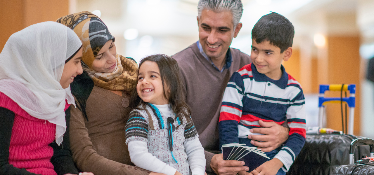 A family of refugees sit at an airport with their luggage next to them and smile.