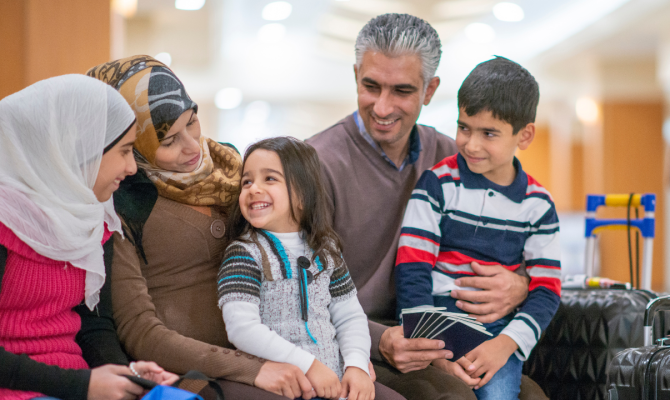 A family of refugees sit at an airport with their luggage next to them and smile.