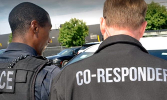 A police officer and a man with the words co-responder on his back look at someone off camera.