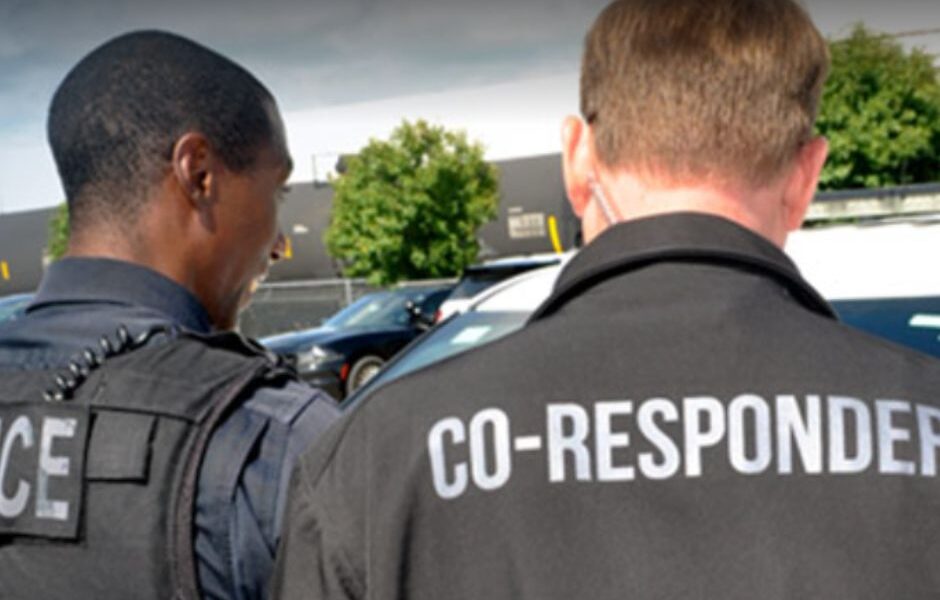 A police officer and a man with the words co-responder on his back look at someone off camera.