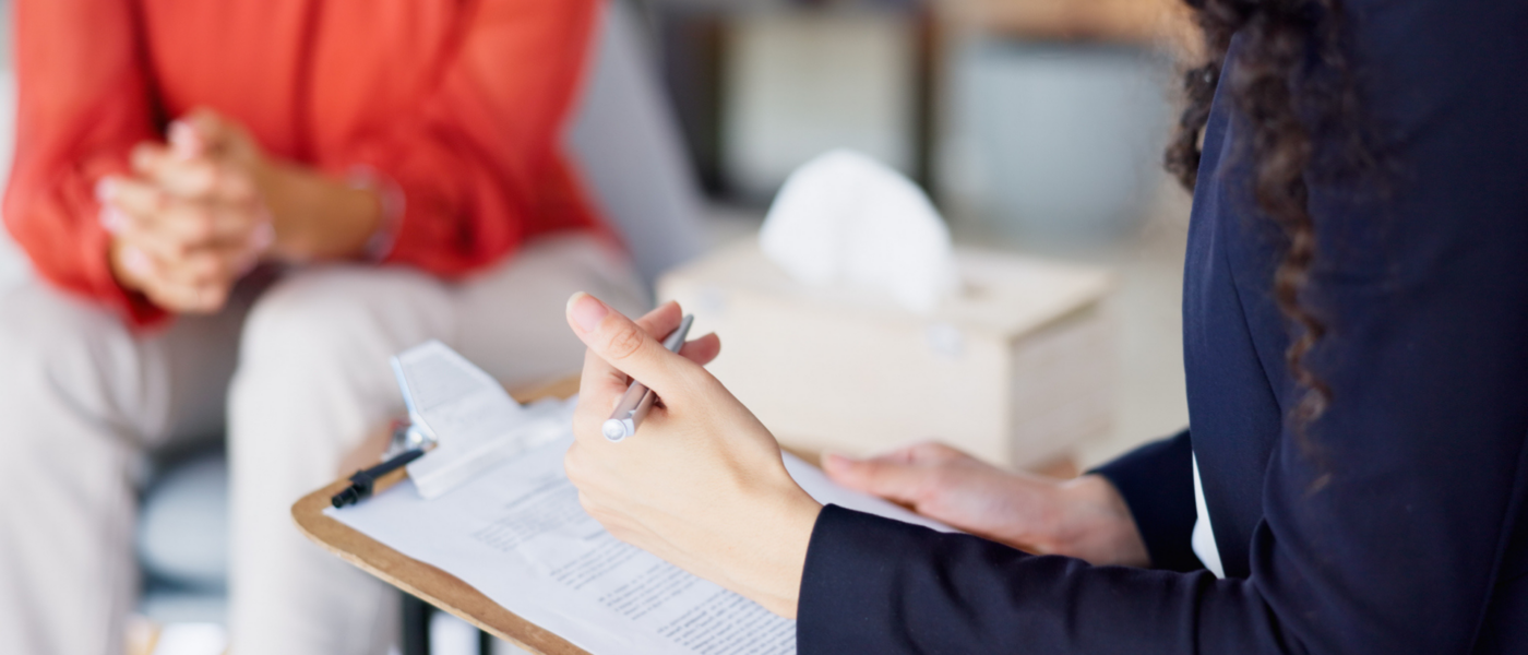 A hand taking notes hovers over a clipboard as a woman in red sits out of focus in the background.