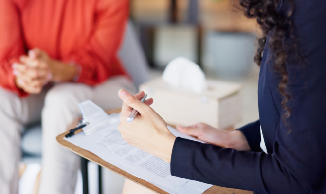 A hand taking notes hovers over a clipboard as a woman in red sits out of focus in the background.