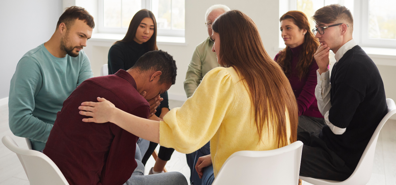 A man holds his face in his hands and is comforted by a woman in a yellow sweater as support group members sit in a small circle.