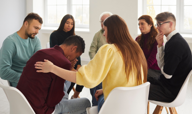 A man holds his face in his hands and is comforted by a woman in a yellow sweater as support group members sit in a small circle.