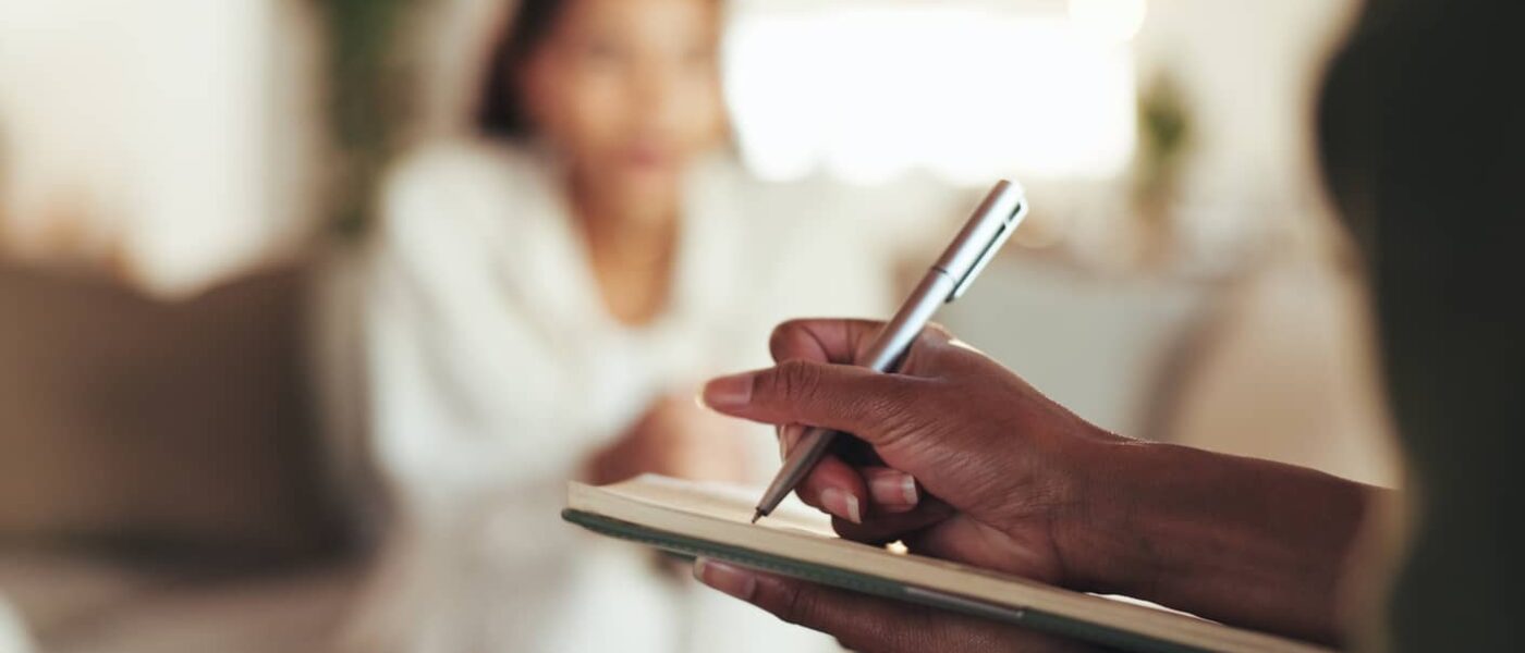 A hand holds over a notepad writing as someone sits out of focus in the background.