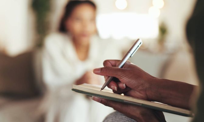A hand holds over a notepad writing as someone sits out of focus in the background.