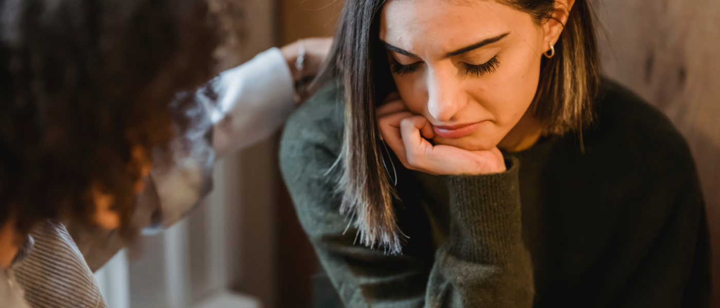 A young woman rests her chin on her hand with her eyes closed and head tilted downward and is comforted by a therapist.