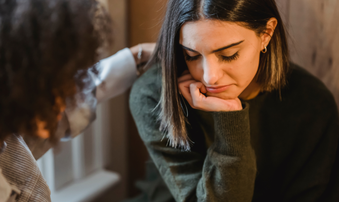 A young woman rests her chin on her hand with her eyes closed and head tilted downward and is comforted by a therapist.