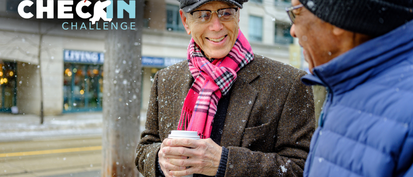 A man in a hat, pink plaid scarf, and wool jacket holds a coffee cup with two hands and smiles at a man in a winter cap and blue coat.