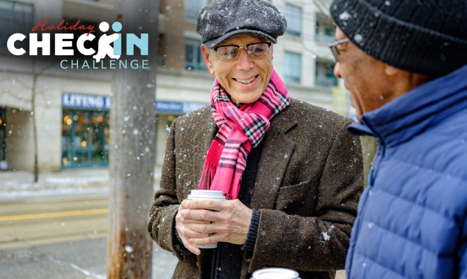 A man in a hat, pink plaid scarf, and wool jacket holds a coffee cup with two hands and smiles at a man in a winter cap and blue coat.