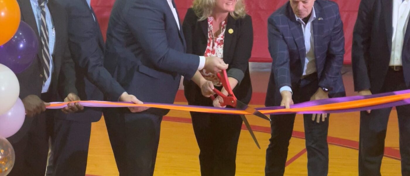 Several people in suits stand in a gym and cut a ribbon with over-sized scissors.