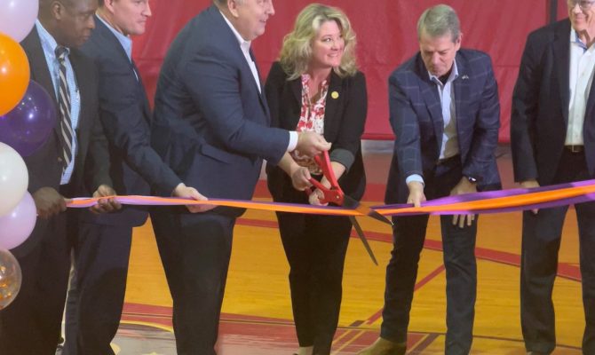 Several people in suits stand in a gym and cut a ribbon with over-sized scissors.
