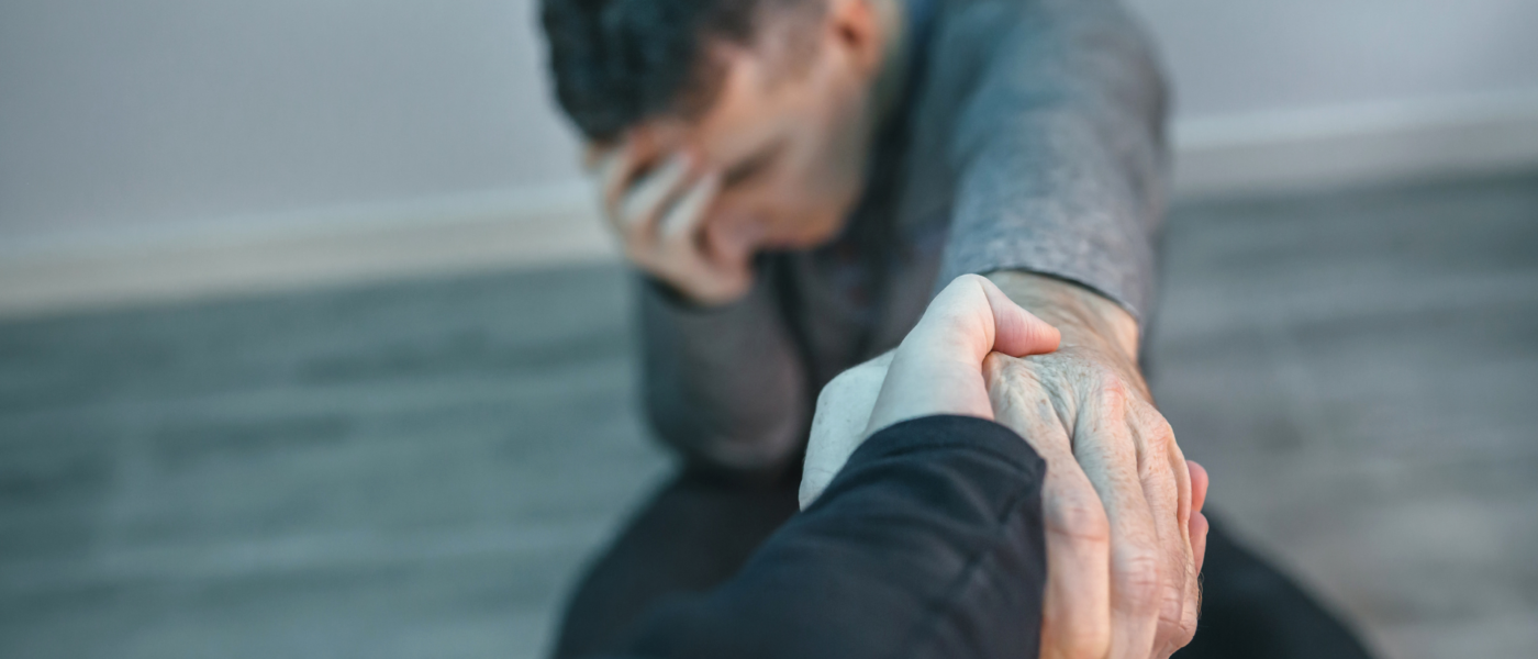A man sits on the floor with his hand on his head eyes closed and outstretches his other arm for assistance off the ground.