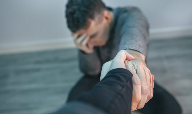A man sits on the floor with his hand on his head eyes closed and outstretches his other arm for assistance off the ground.