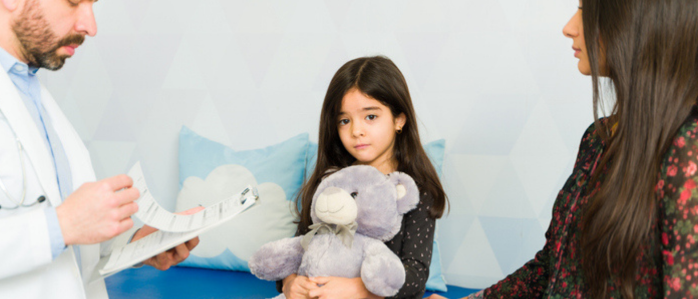 A doctor looks at his clipboard as a child peers sadly while holding a large gray teddy bear and her mother sits beside her facing the doctor.