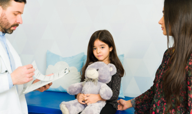 A doctor looks at his clipboard as a child peers sadly while holding a large gray teddy bear and her mother sits beside her facing the doctor.