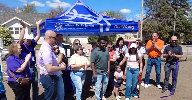 A group of people stand in front of a tent for Lutheran Family Services and smile as a ribbon has just been cut in a community garden.