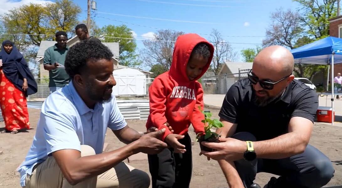 A small child wearing a red hoodie plants a vegetable with the assistance of two men on each side.