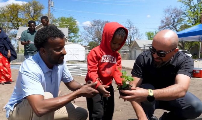 A small child wearing a red hoodie plants a vegetable with the assistance of two men on each side.