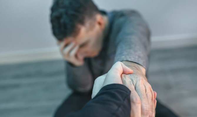 A man sits on the floor with his hand on his head eyes closed and outstretches his other arm for assistance off the ground.