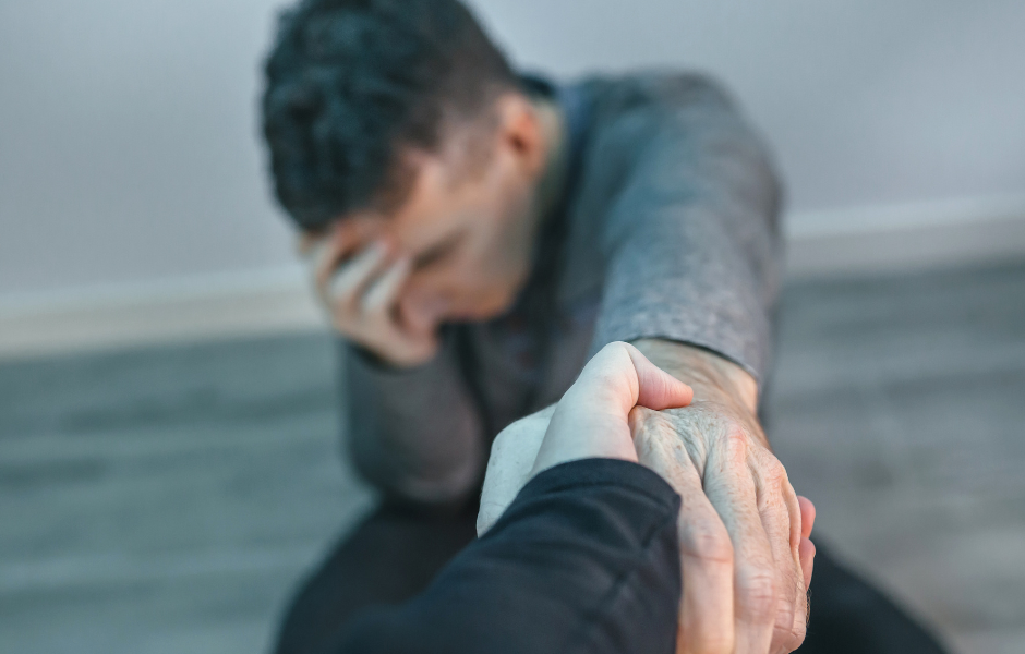 A man sits on the floor with his hand on his head eyes closed and outstretches his other arm for assistance off the ground.