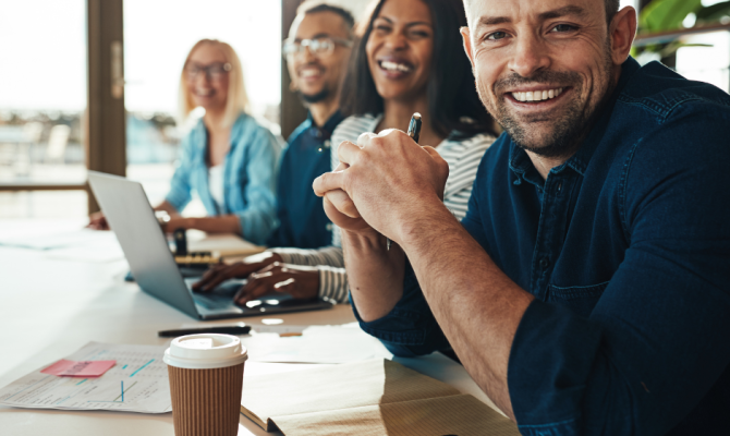 A Man sits behind a table with his hands clasped smiling in your direction several out of focus co-workers smiling down the table.