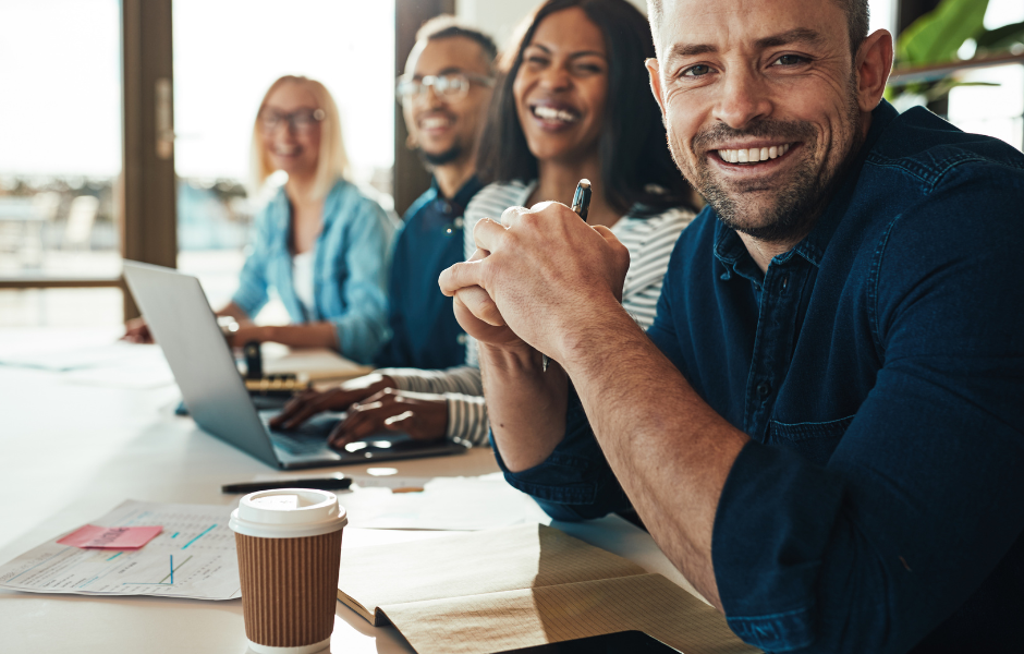 A Man sits behind a table with his hands clasped smiling in your direction several out of focus co-workers smiling down the table.