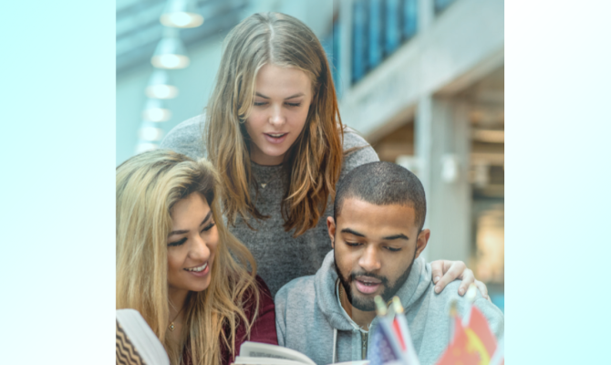 A young woman looks over the shoulders of two studying students to read a book.