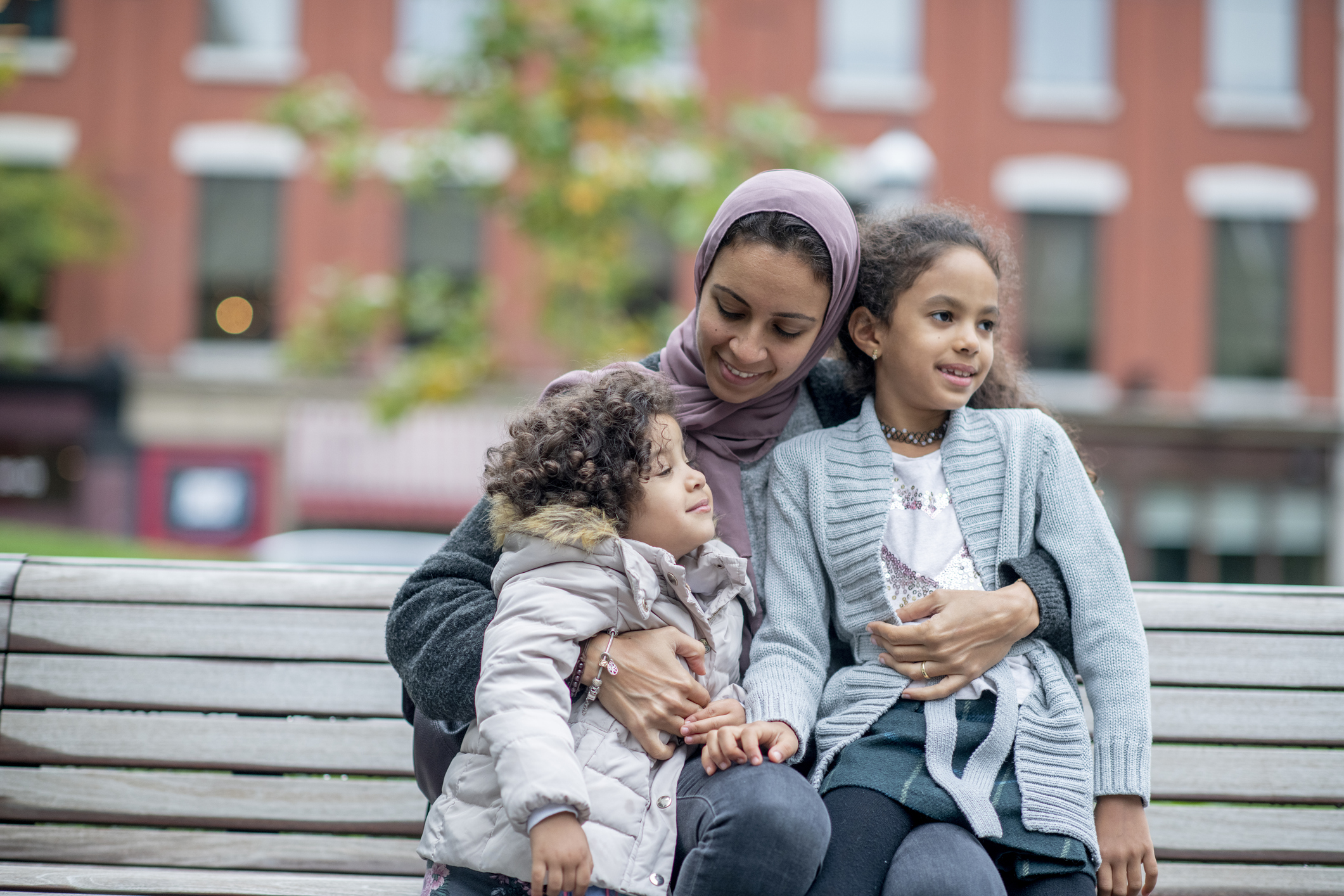 A refugee mother hugs her daughters on a park bench in their new community,