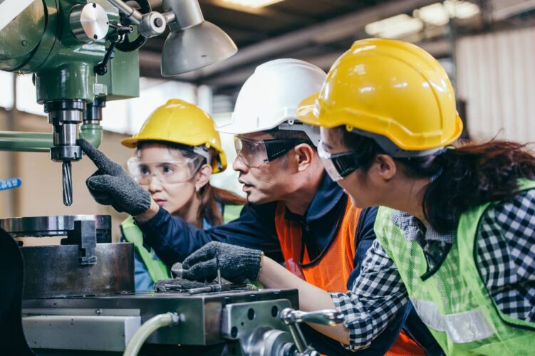 An instructor wearing a hard hat shows two students in hard hats a feature of a lathe.