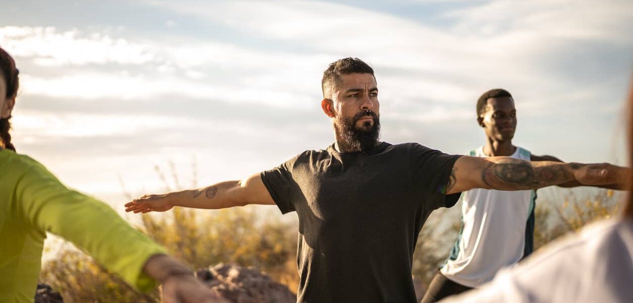 A bearded man performs yoga in a field along with others in a class.