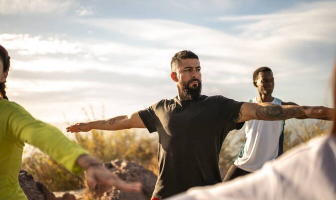 A bearded man performs yoga in a field along with others in a class.