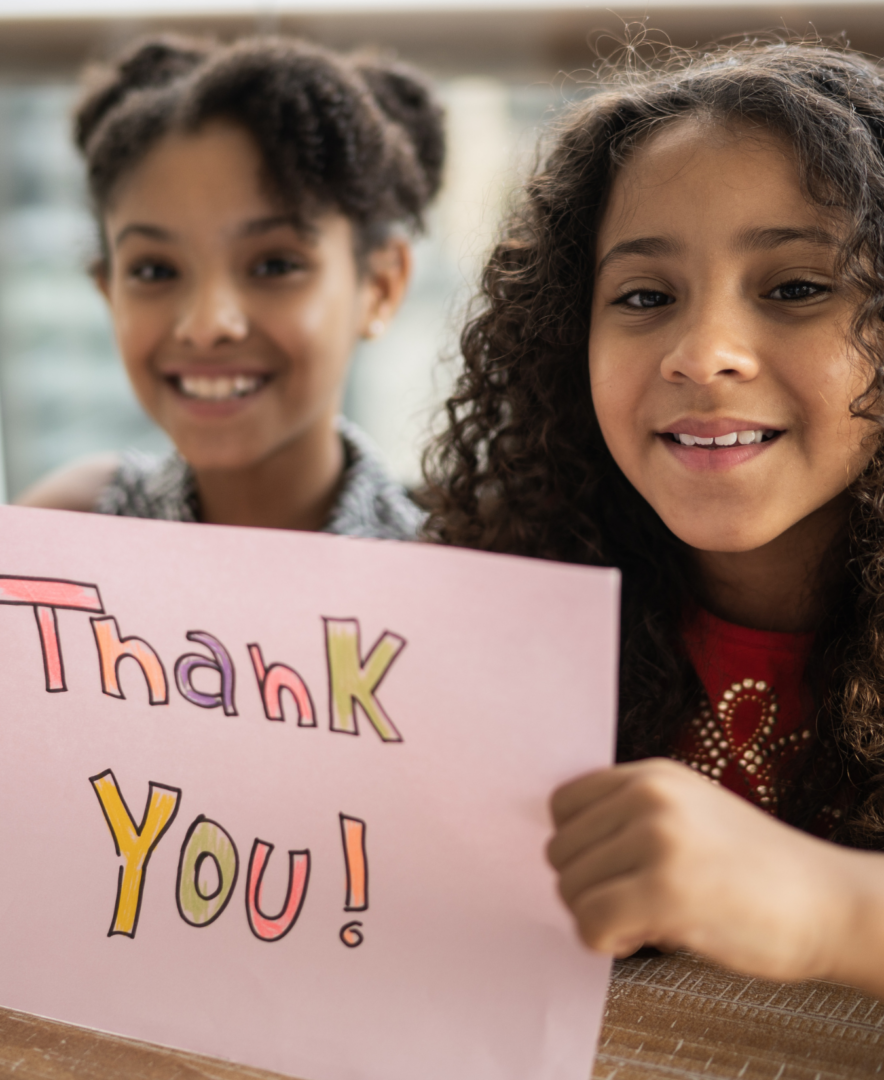 Two girls hold a hand-made, crown- colored sign that says "thank you."