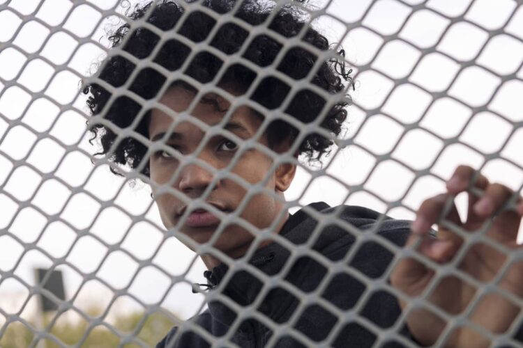 A young man looks through a chain-link fence to the distance.