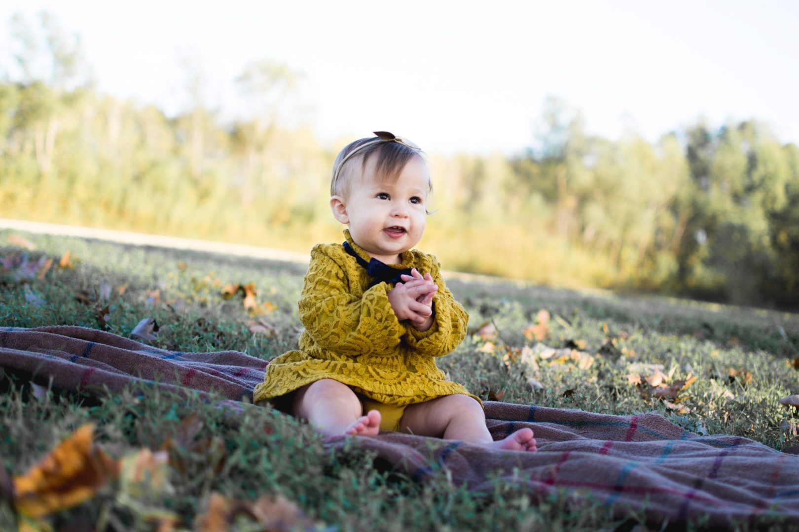 A girl clasps her hands together and smiles as she sits on a blanket on a slight hill in a green field littered sporadically with fallen leaves.
