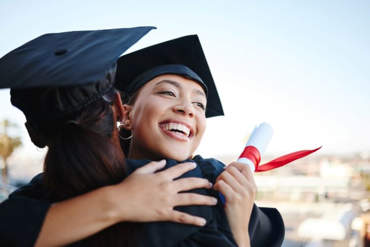 A college graduate in cap and gown embraces a fellow graduate as she holds a diploma in her hand.