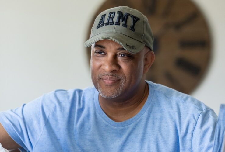 An army veteran wearing a blue shirt and ARMY hat sits and listens to someone speak.